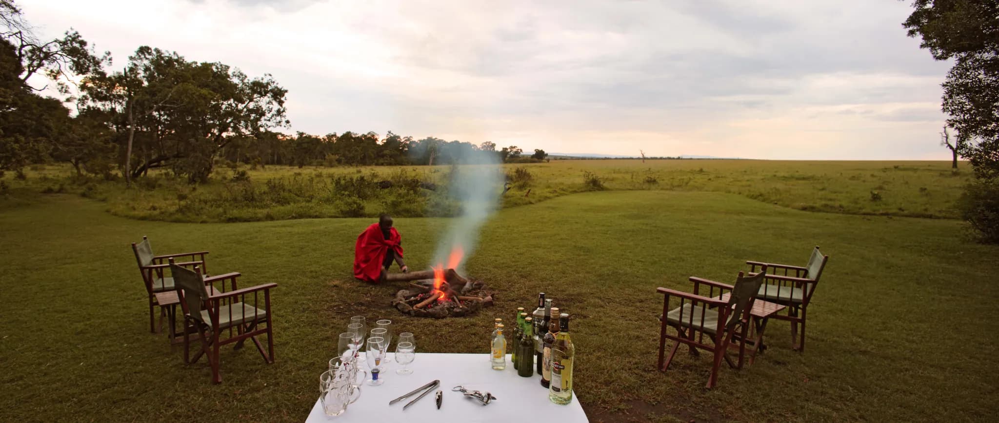 A Maasai guide tends the open campfire at dusk at Elephant Pepper Camp, the plains stretching beyond — Photo: Elewana Collection