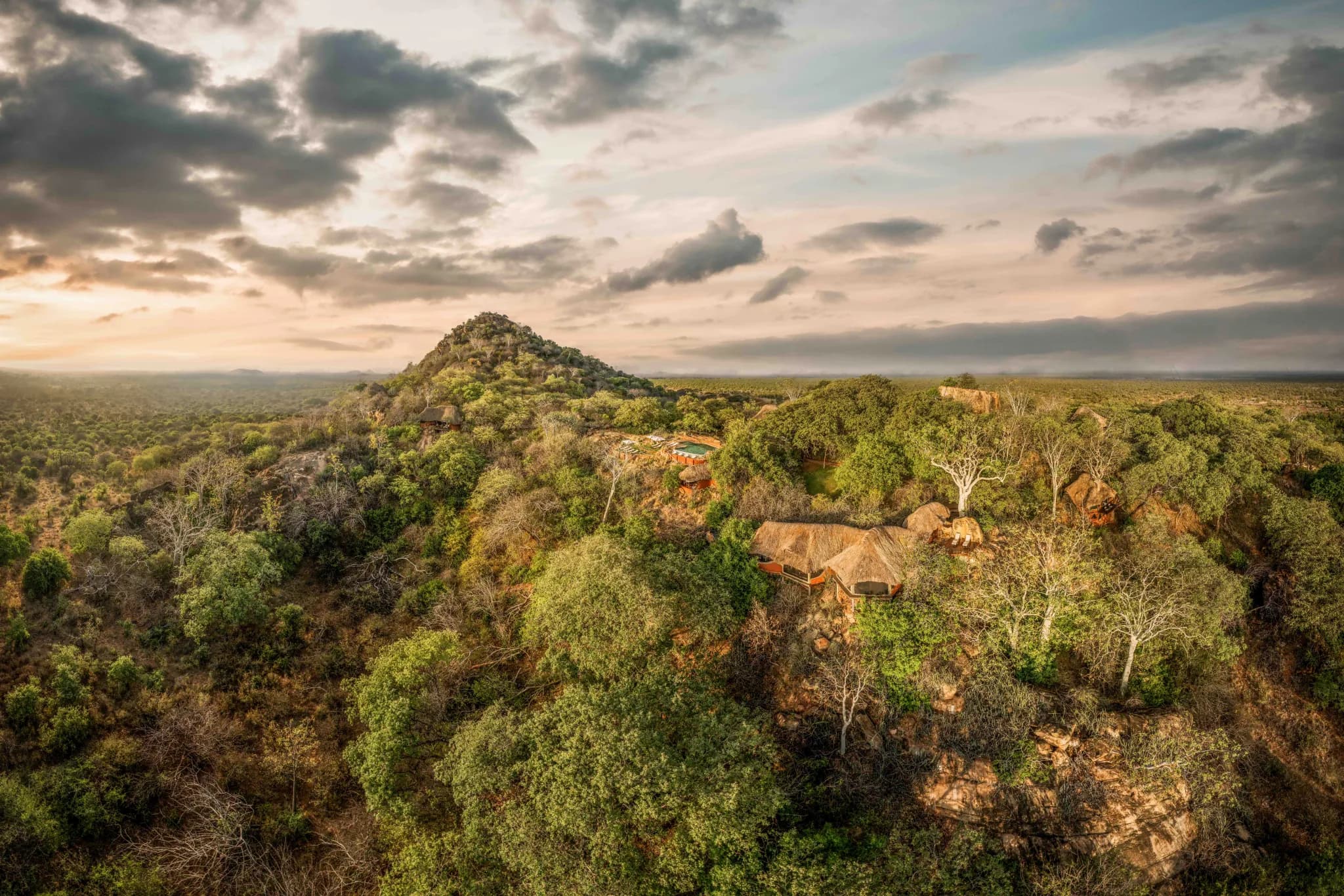 Aerial view of Elsa's Kopje woven into the rock and vegetation of Mughwango Hill, the plains beyond — Photo: Elewana Collection