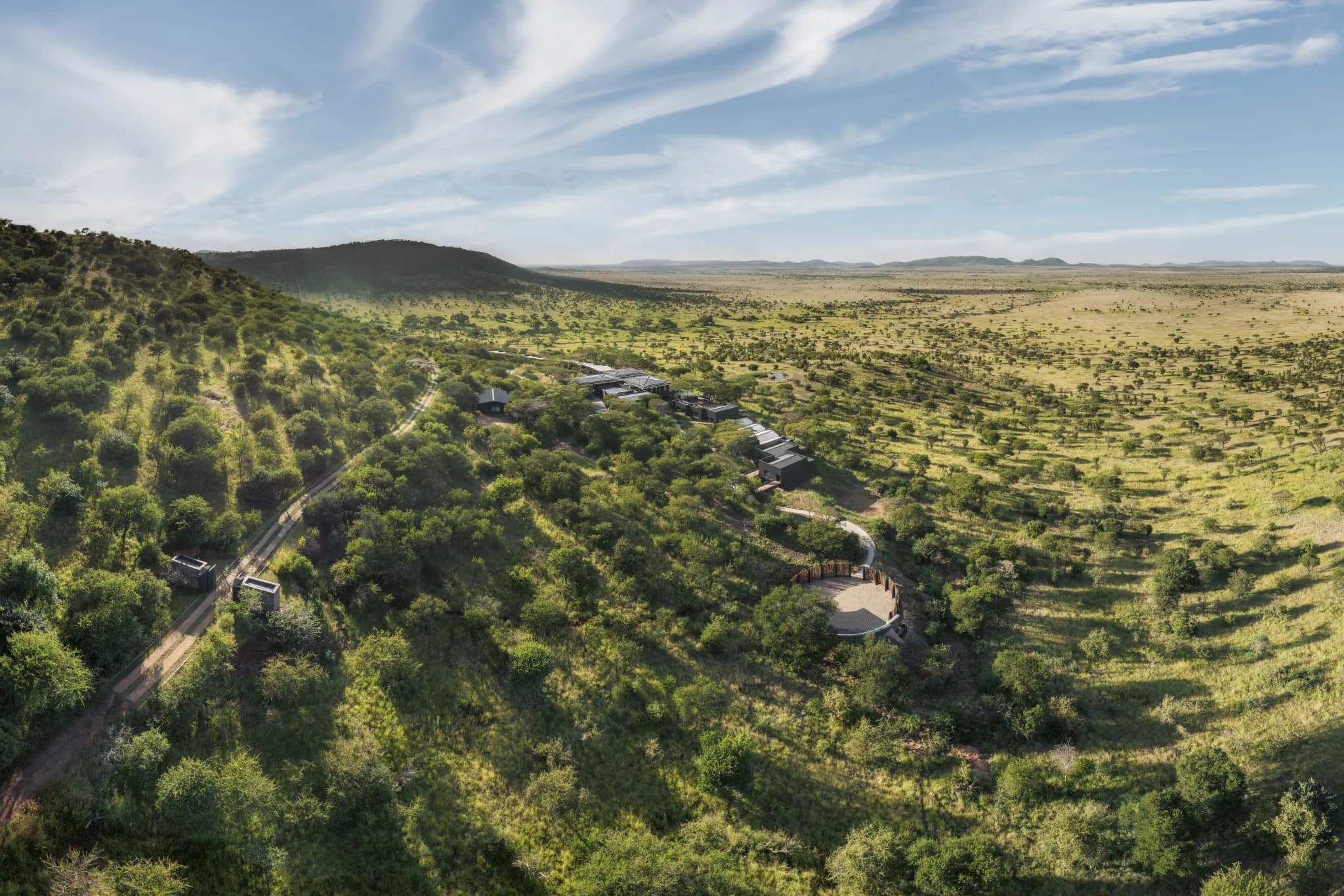 Aerial view of a safari lodge set into wooded hills above the open Serengeti plains stretching to the horizon